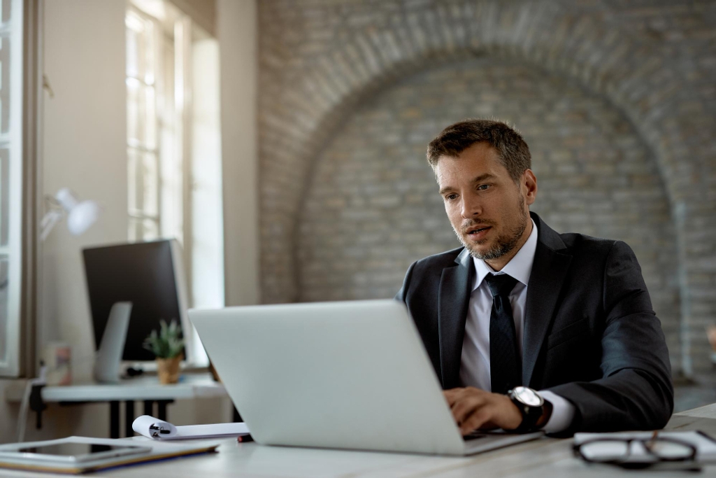 A Professional Man in a Dark Suit Sits at a Desk, Focused on His Laptop in a Modern Office With Exposed Brick Walls and Soft Daylight. Documents, a Tablet and Glasses Rest on the Desk, Suggesting He Is Revie